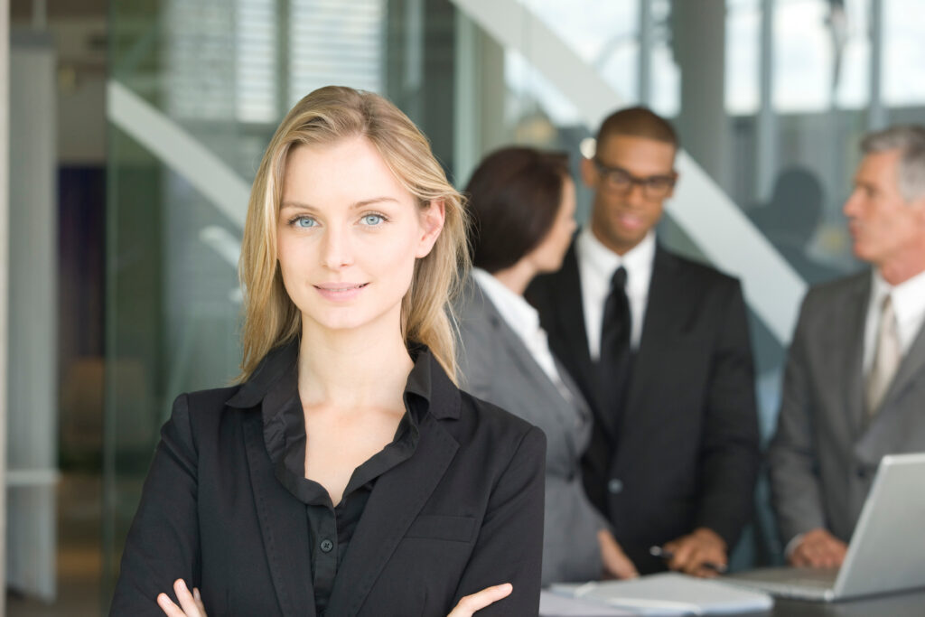 A confident sales professional standing in a boardroom with her colleagues in the background.